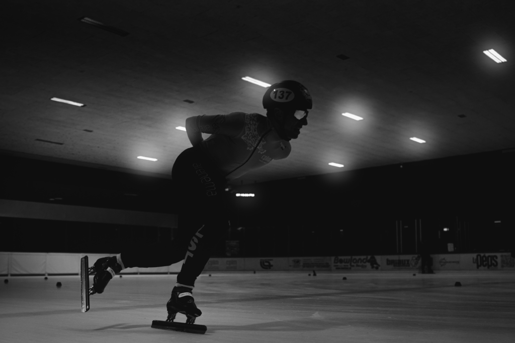 Patineur de short track en pleine poussée, vu au ras de la glace en noir et blanc à la Vaudoise Arena de Lausanne.