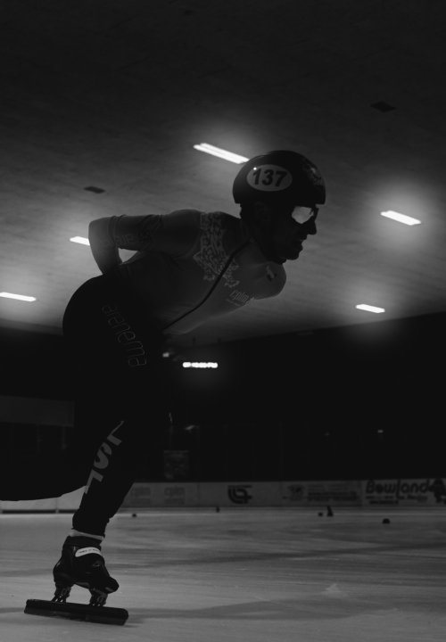 Patineur de short track en pleine poussée, vu au ras de la glace en noir et blanc à la Vaudoise Arena de Lausanne.
